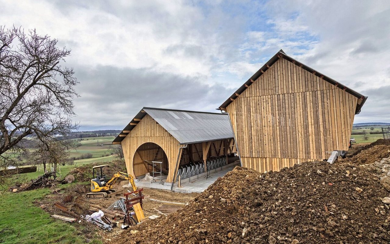 Zwei fast fertige Stallgebäude aus Holz sehen an einem sanften Hügel. Das eine Gebäude hat ein rundes Tor.