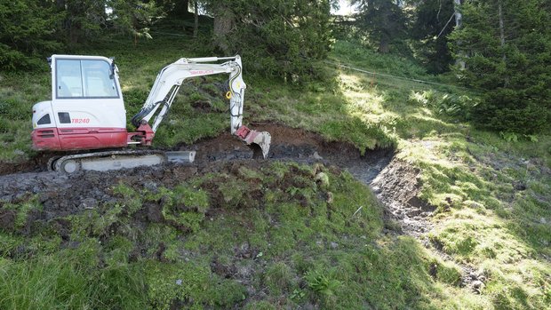 Wasserversorgung Alpwirtschaft Ein Bagger steht im Hang, daneben gluckert ein kleines Wasserrinnsal hinunter.