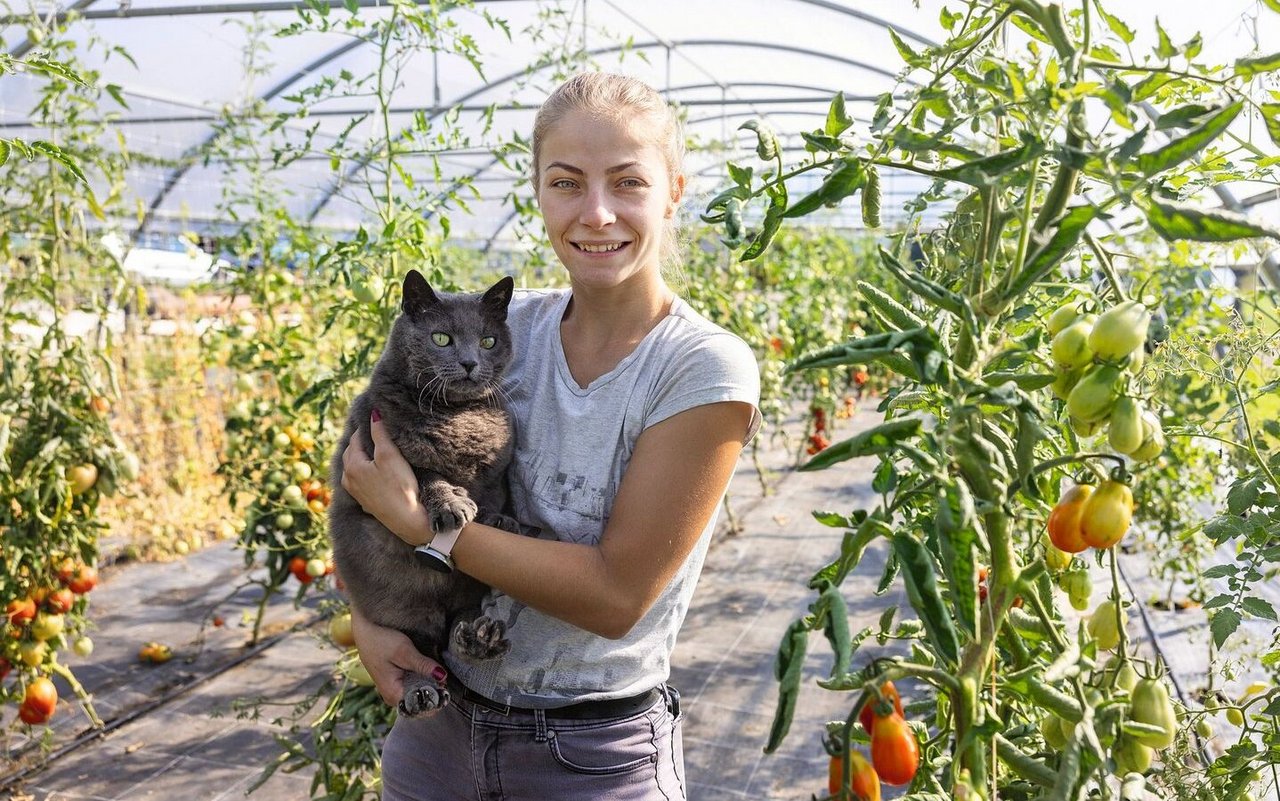 Porträt von Kati Fábián, die im Gewächshaus steht und einen grauen Kater auf dem Arm trägt.