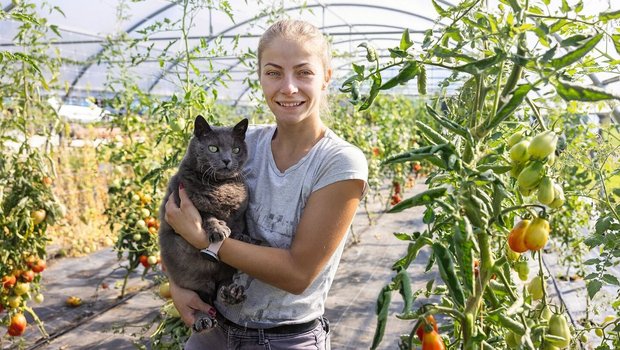 Porträt von Kati Fábián, die im Gewächshaus steht und einen grauen Kater auf dem Arm trägt.