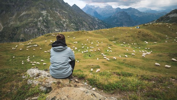 Eine Frau sitzt erhöht auf einem Stein und beobachtet die Schafherde auf der Alpweide unter ihr.