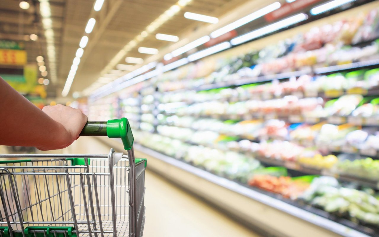 Eine Hand schiebt einen leeren Einkaufswagen durch die Regale eines Supermarkts.