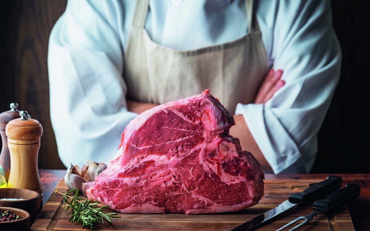 T-Bone Steak auf olzbrett, dahinter Metzger mit verschränkten Armen..