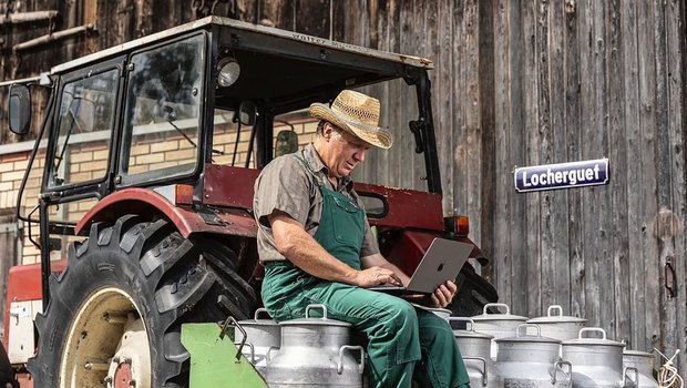 Landwirt sitzt auf Milchkannen und schreibt auf einem Notebook.