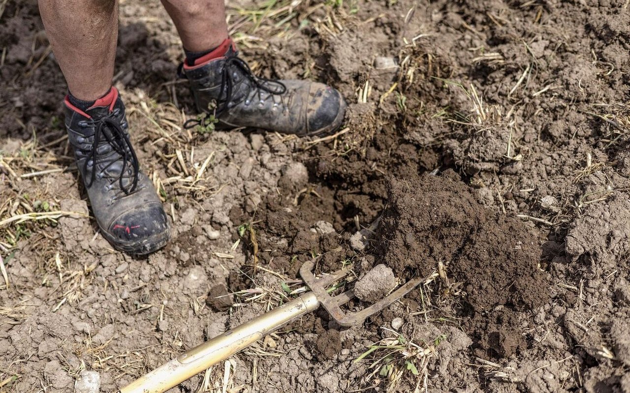 Auf dem unbedeckten Boden liegt eine Spatengabel, daneben sind zwei paar Füsse in Arbeitsschuhen zu sehen.