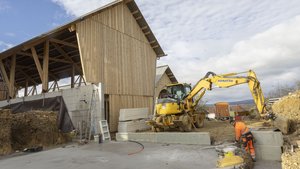 Vor einem fast fertigen Stallgebäude steht ein gelber Bagger.