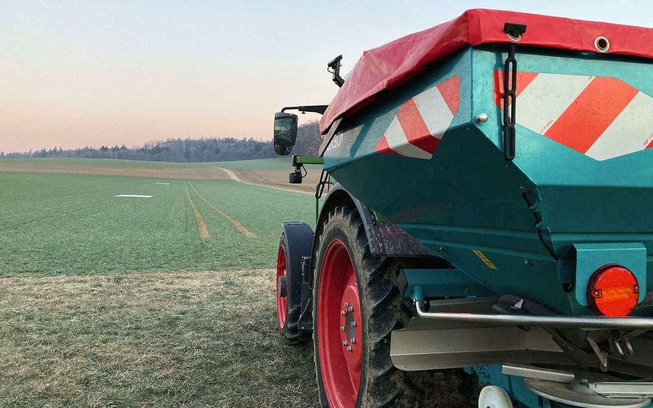 ein Traktor mit angehängtem Düngerstreuer steht am Rand eines Weizenfelds.